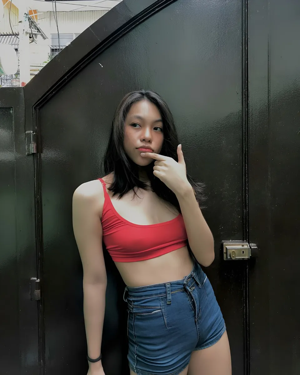 Abegail Bennett in red top and denim shorts leaning against dark door