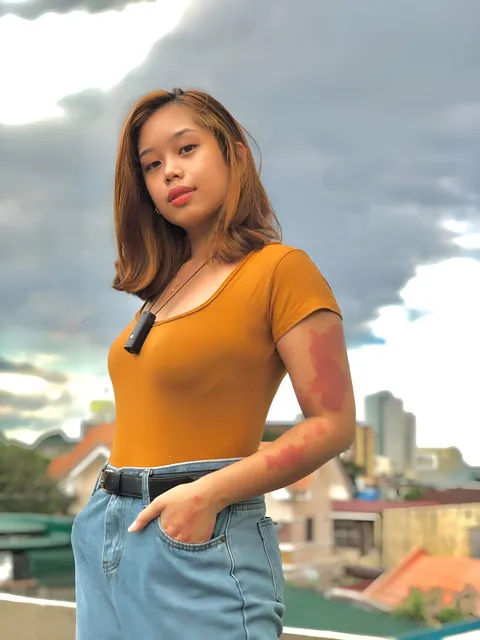 Ally Romano in orange top and jeans stands on rooftop with cloudy sky
