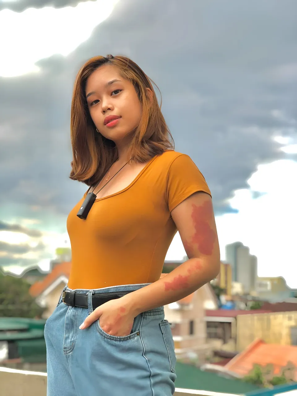 Ally Romano in orange top and jeans stands on rooftop with cloudy sky
