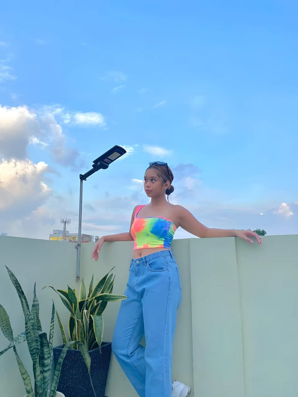 Ally Romano in colorful top and jeans leans against a wall on a rooftop
