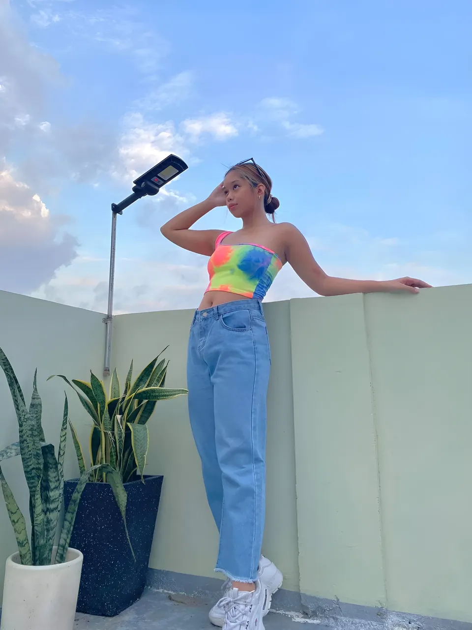 Ally Romano in tie-dye top and jeans stands on rooftop with plants