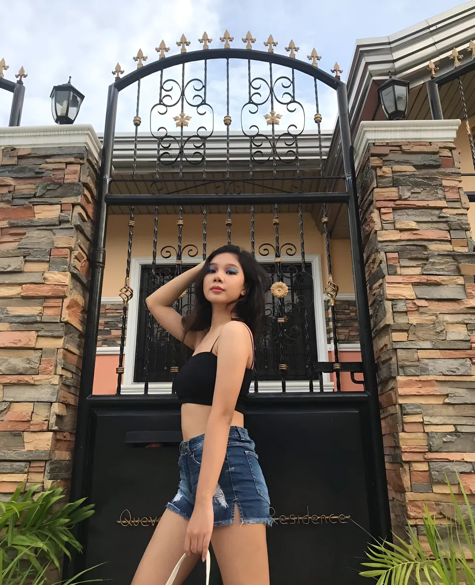 Ariane Quevedo in black top and denim shorts stands by ornate gate