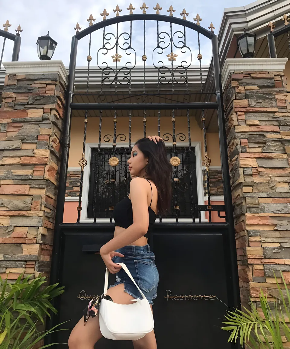 Ariane Quevedo in black top and denim shorts stands beside black metal gate