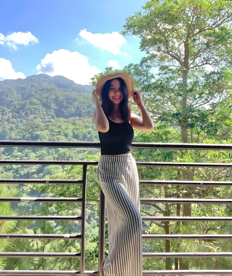 Ariane Quevedo wearing striped pants and a black top holds a hat on a balcony