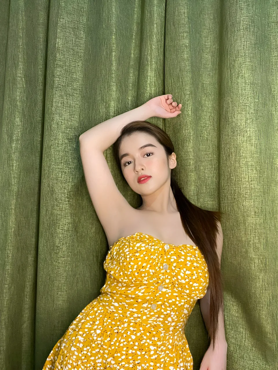 Asia Catacutan wearing a yellow floral dress raises one arm against a green curtain