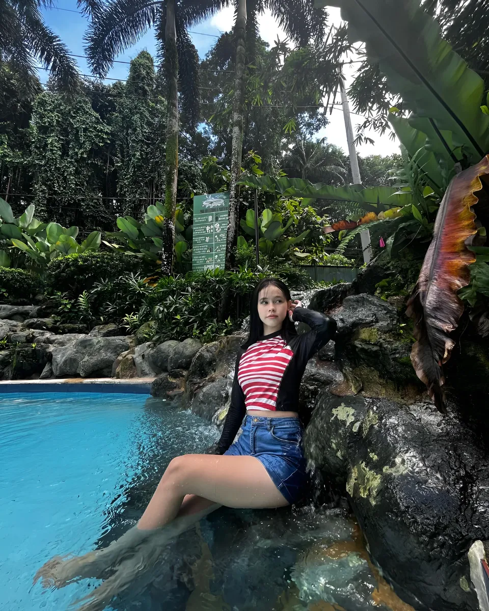 Ayesha Oprenario in striped top sits in pool beside rocks and foliage