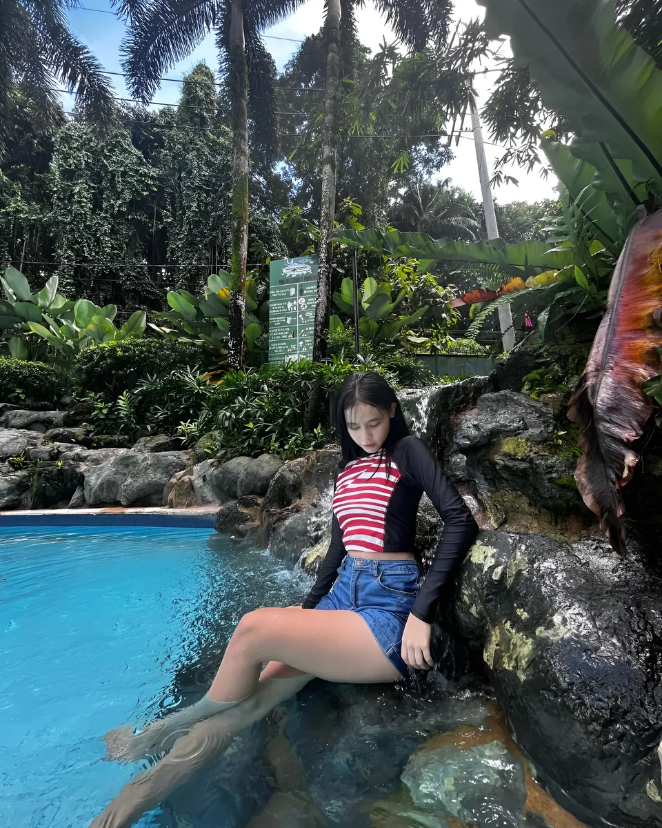 Ayesha Oprenario in striped top sits in pool beside rocky waterfall feature