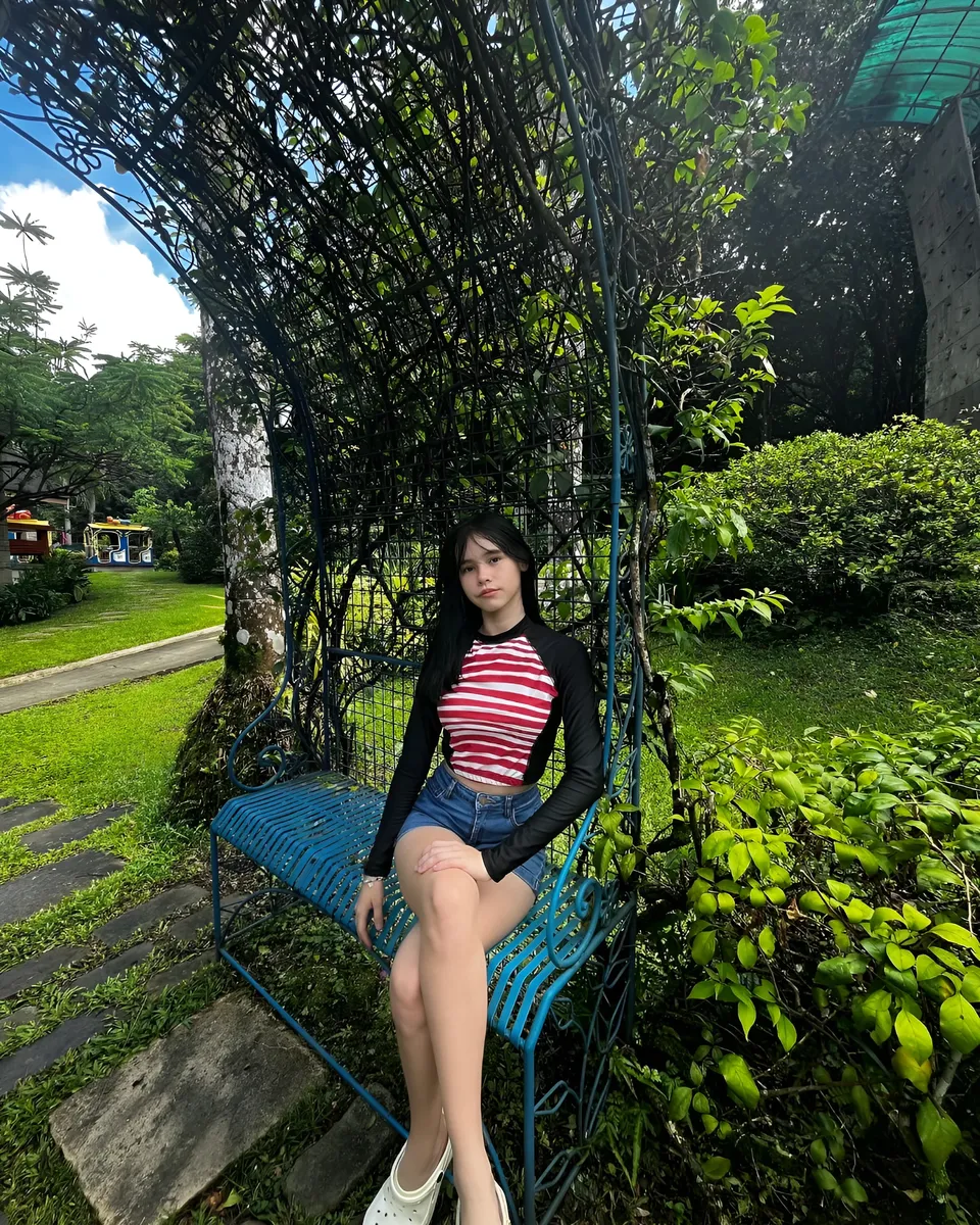 Ayesha Oprenario sits on blue bench under green trellis in garden