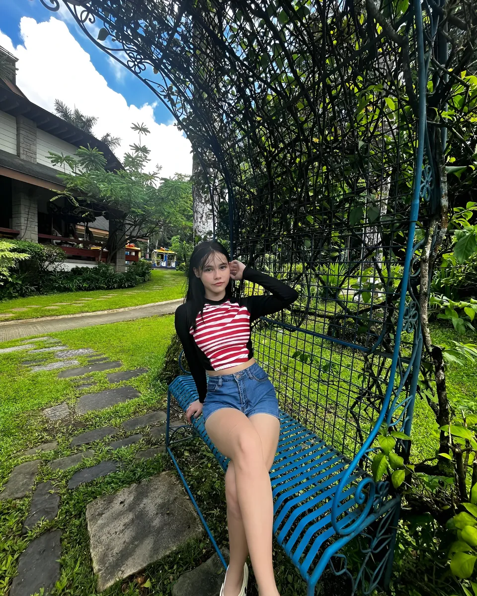 Ayesha Oprenario sits on blue bench outdoors near greenery and building