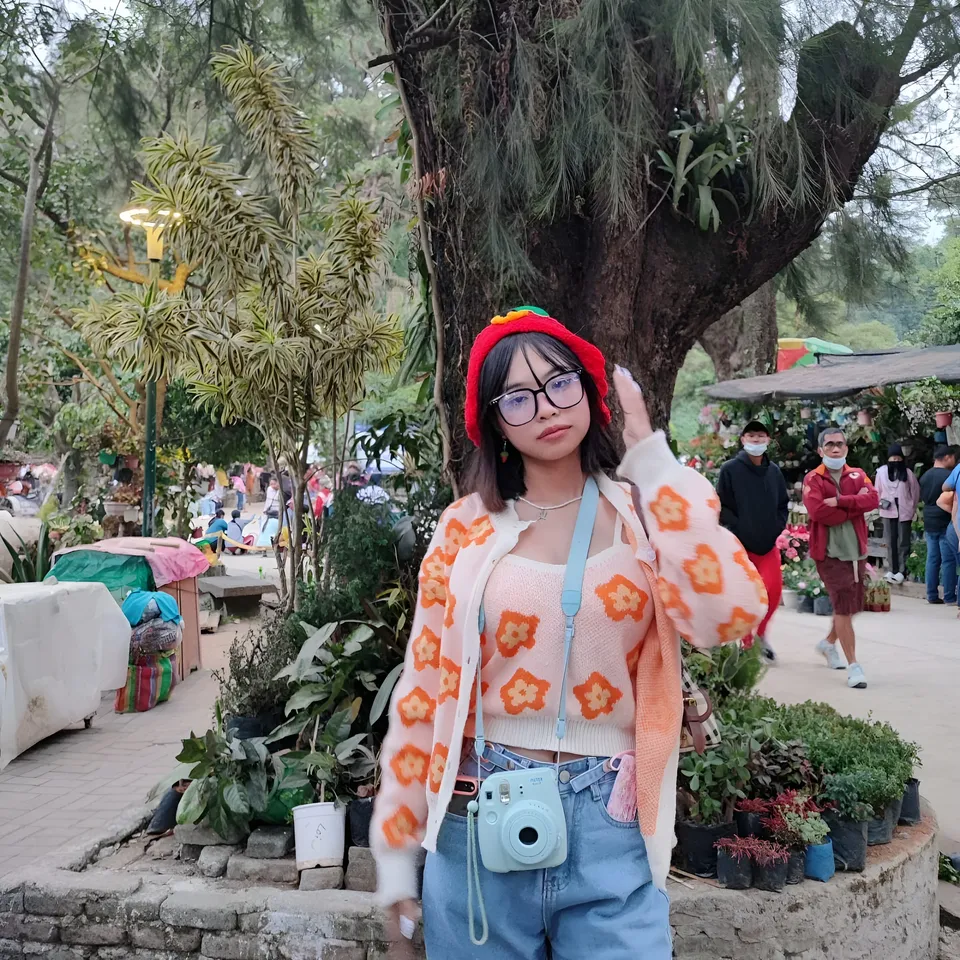 Beri Sinclair in patterned cardigan and red hat stands outdoors near plants