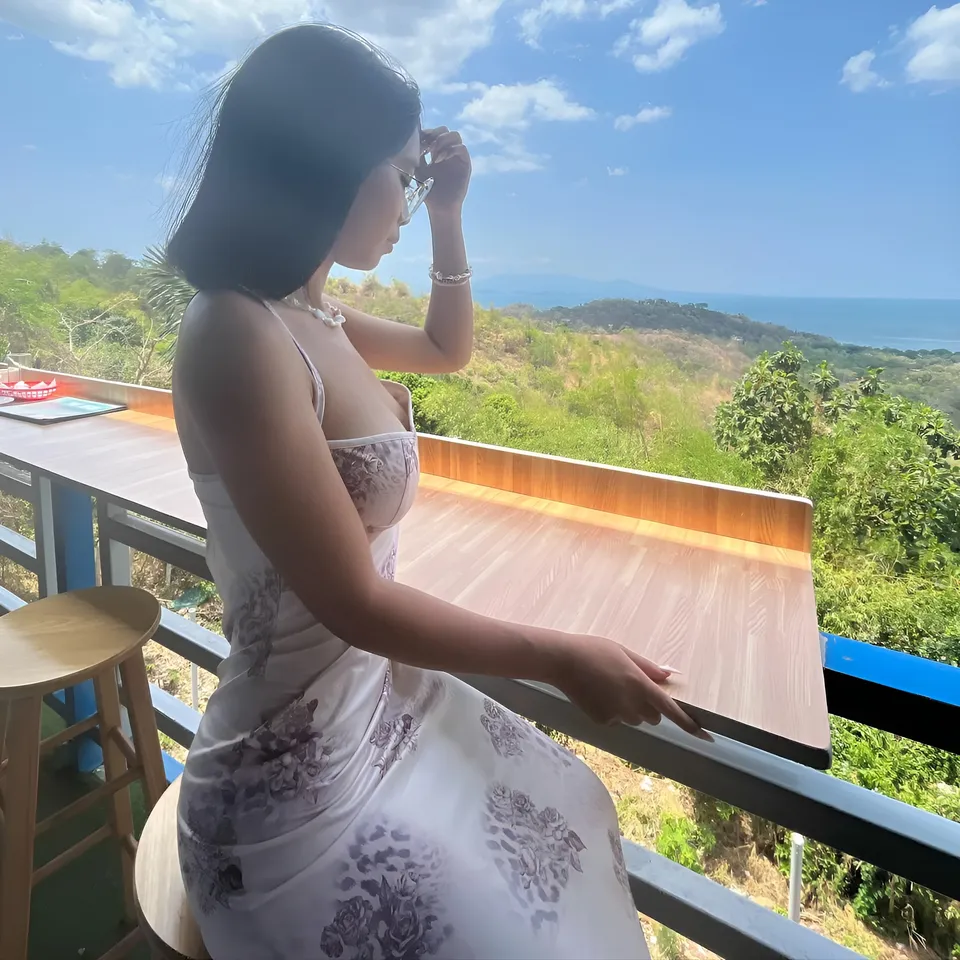 Beri Sinclair in floral dress seated on balcony overlooking green hills and sea