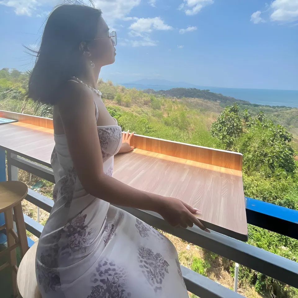 Beri Sinclair in floral dress seated on balcony overlooking green hills and sea