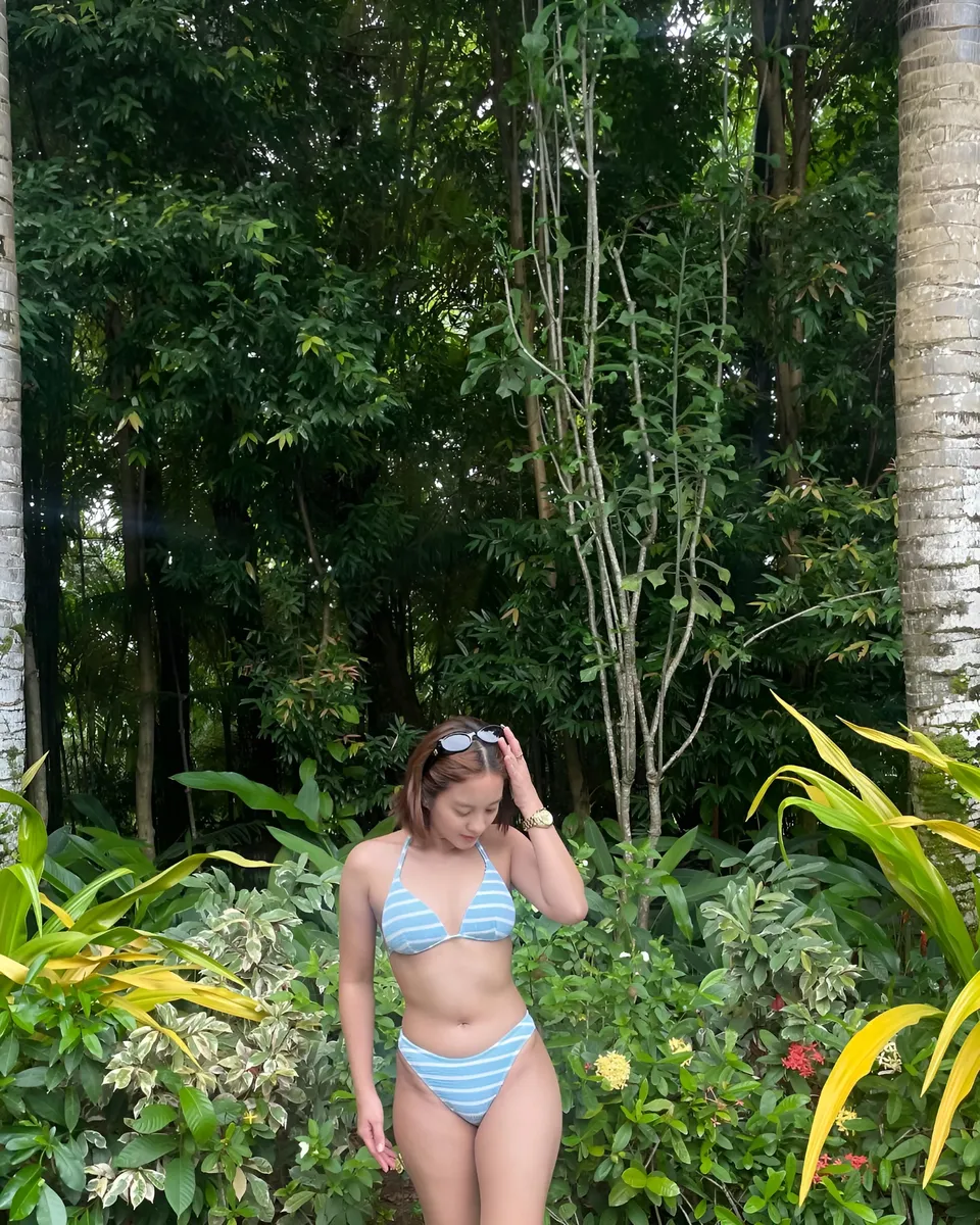 Cassidy Rowan in striped bikini stands among lush green foliage and plants