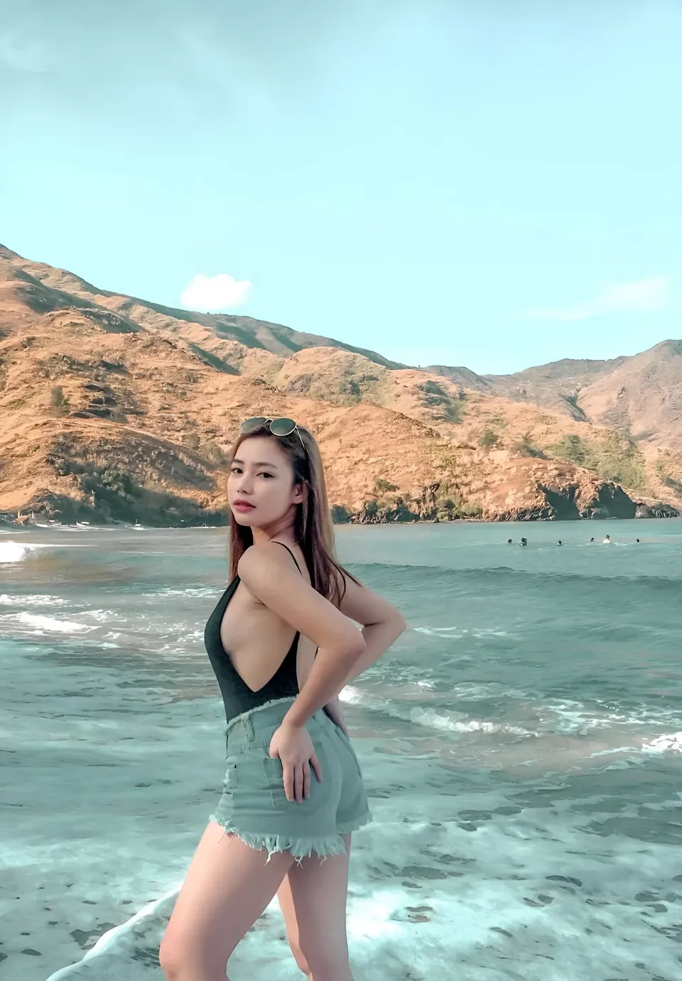 Charlene Migalang in black top and denim shorts stands at ocean's edge with mountains behind