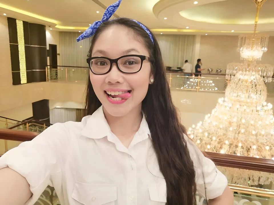 Chloe Arden wearing glasses and a white shirt takes a selfie beside a chandelier