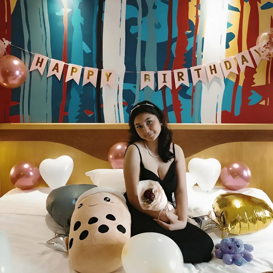 Danisha Vale in black dress seated on bed with balloons and banner
