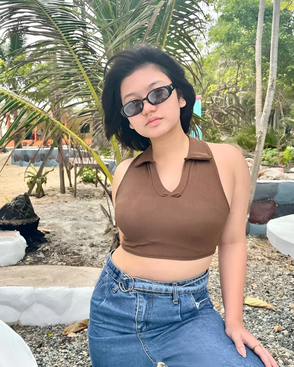 Ellidiethe Rielle wearing brown top and jeans, seated outdoors near palm trees