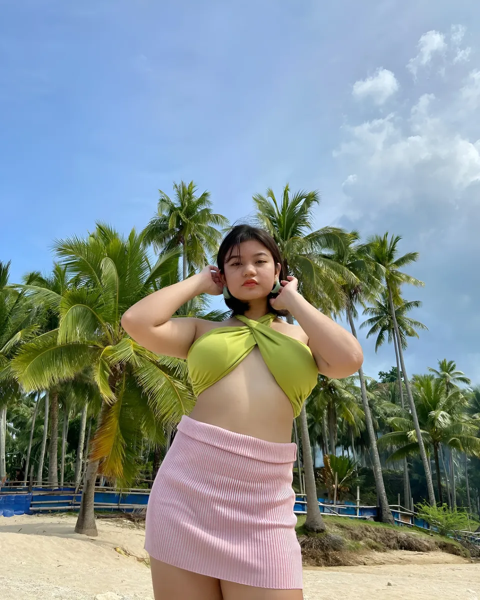Ellidiethe Rielle in green top and pink skirt on sandy beach with palm trees