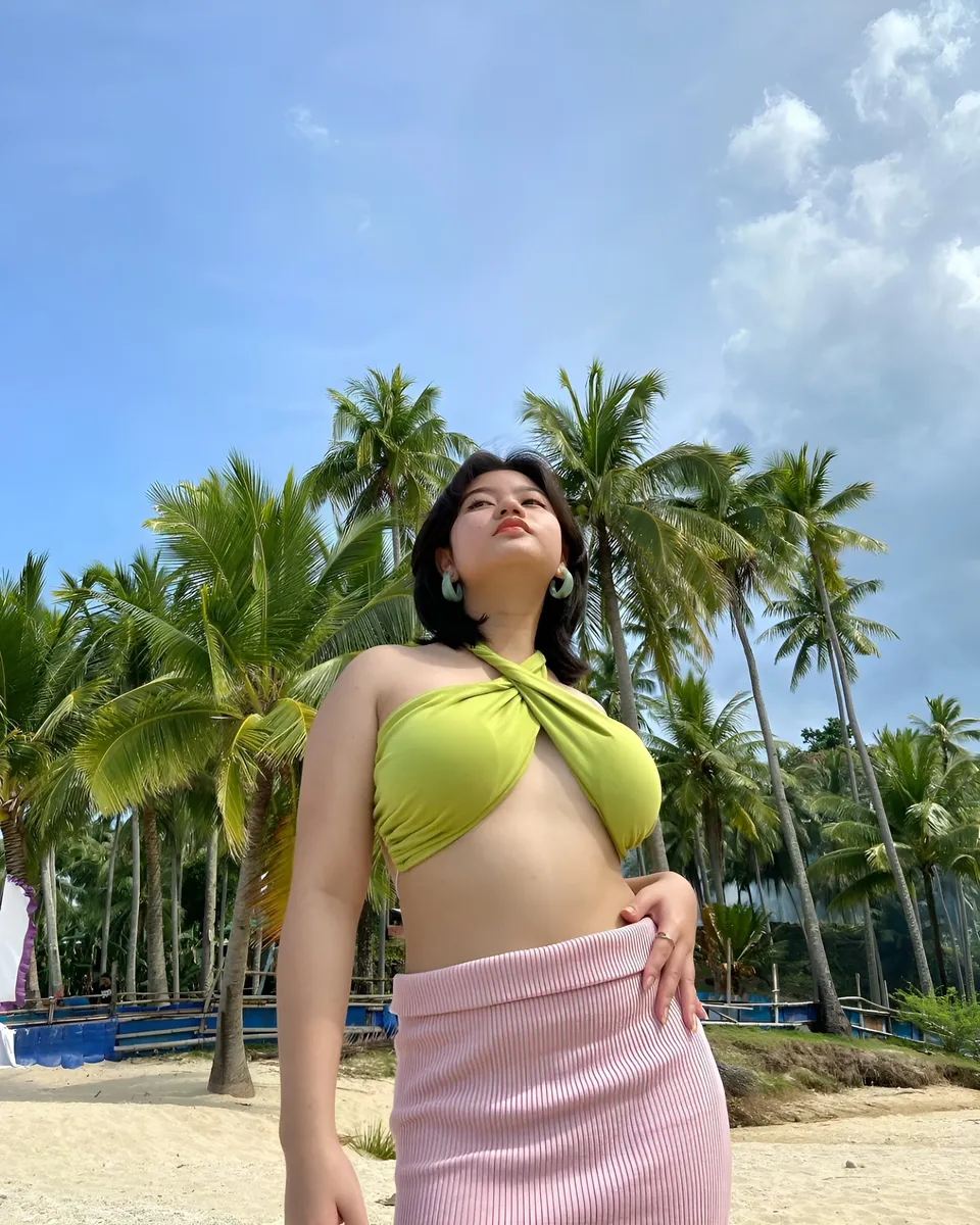 Ellidiethe Rielle in green top and pink skirt stands on sandy beach with palm trees
