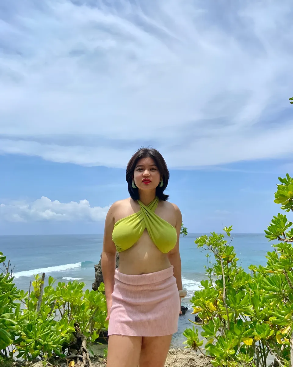 Ellidiethe Rielle in green top and pink skirt stands on beach with ocean and sky