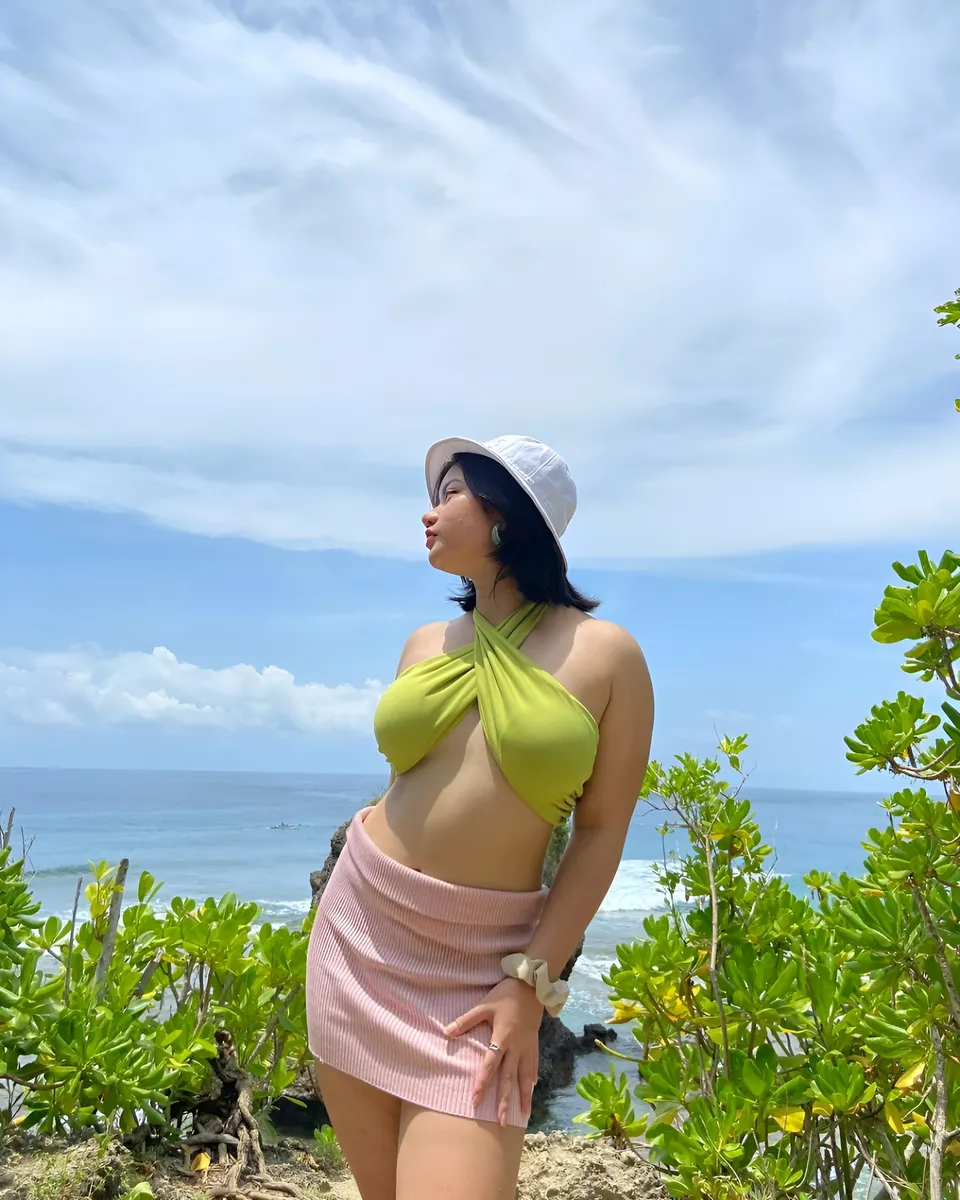 Ellidiethe Rielle in green top and pink skirt stands near ocean with green foliage