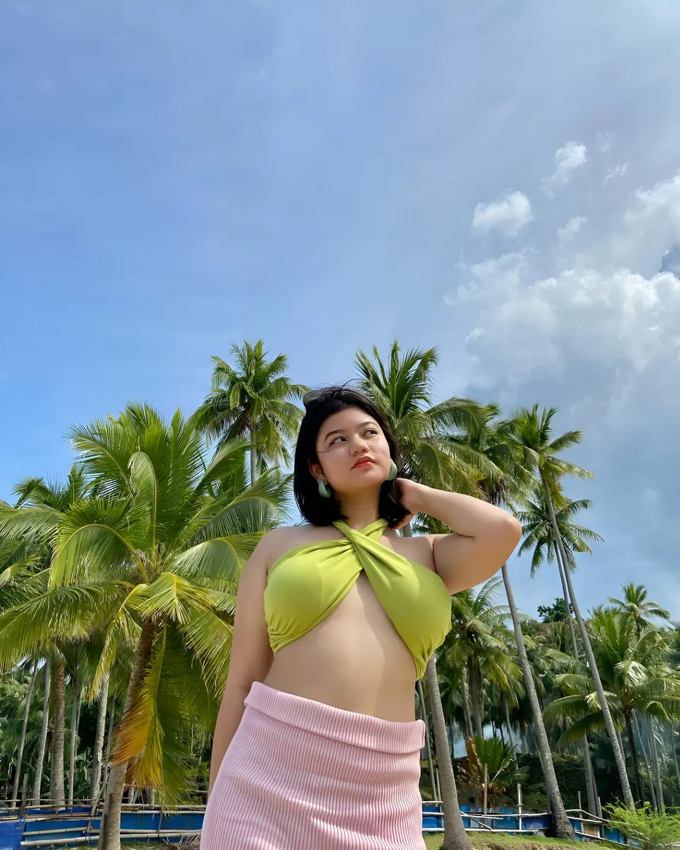 Ellidiethe Rielle in green top and pink skirt stands among palm trees outdoors