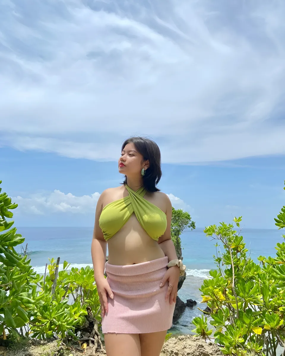 Ellidiethe Rielle in green top and pink skirt stands on cliff overlooking ocean