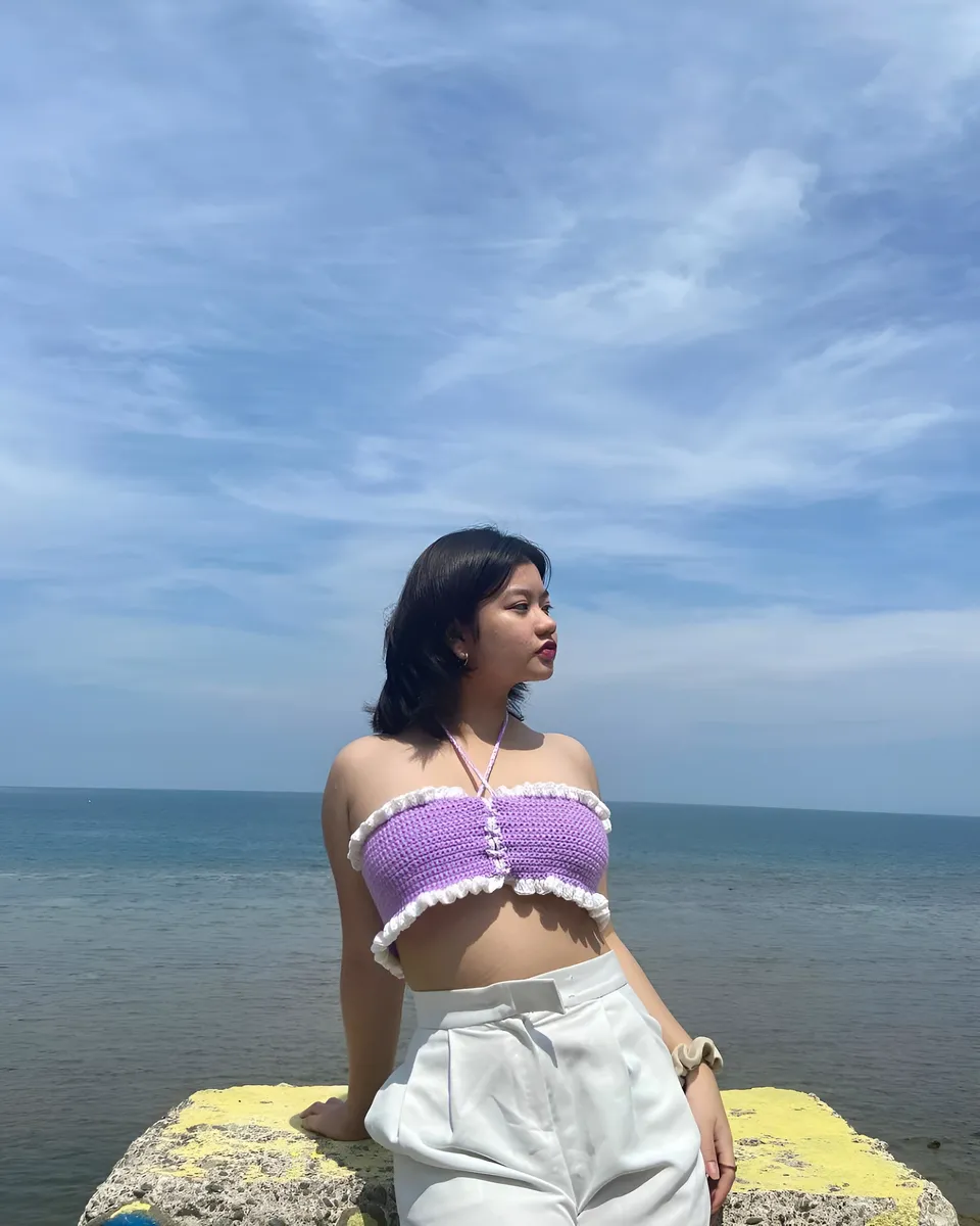 Ellidiethe Rielle in purple crocheted top and white pants stands by sea