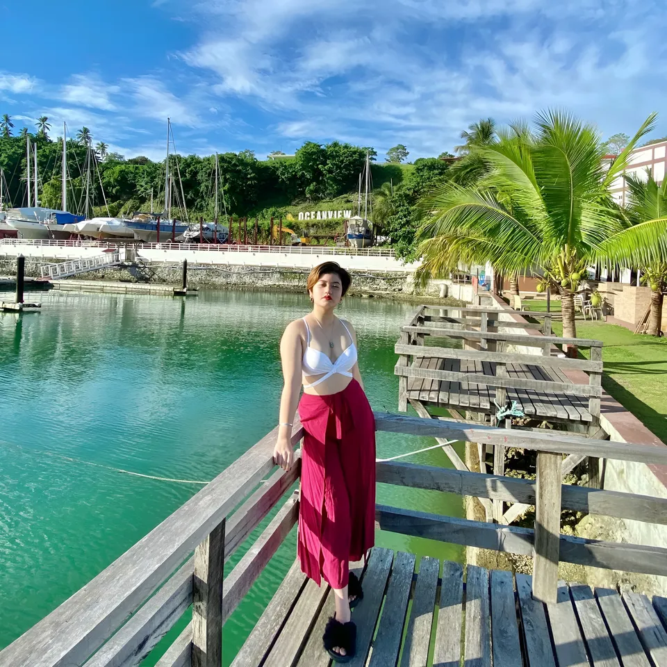 Ellidiethe Rielle in white top and red wrap skirt stands on wooden dock near water