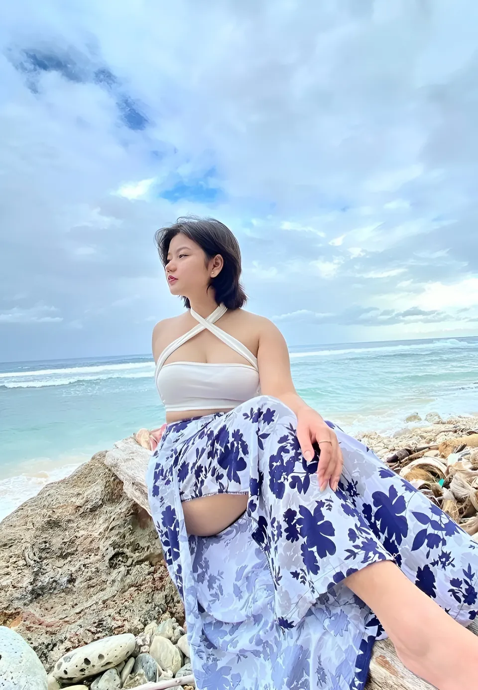 Ellidiethe Rielle in white top and floral pants sits on rocks by ocean