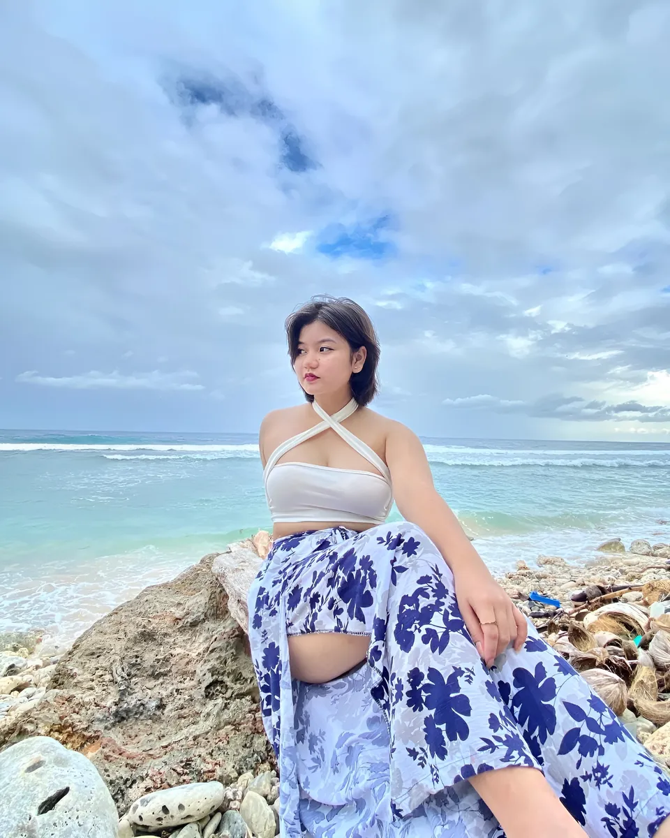 Ellidiethe Rielle in white top and floral pants sits on rock by ocean