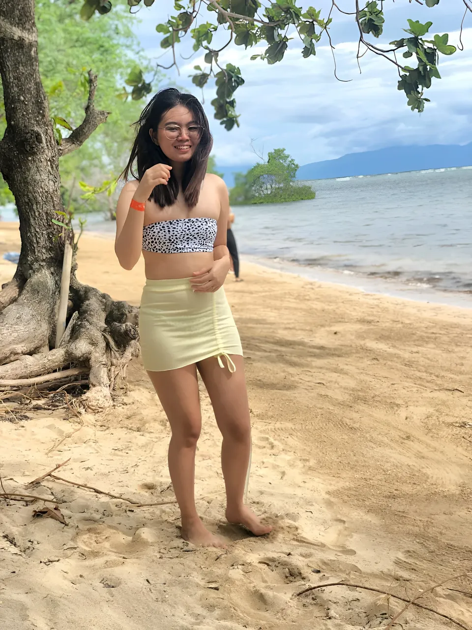 Erica Kyle wearing a patterned top and light skirt stands on a beach beside a tree