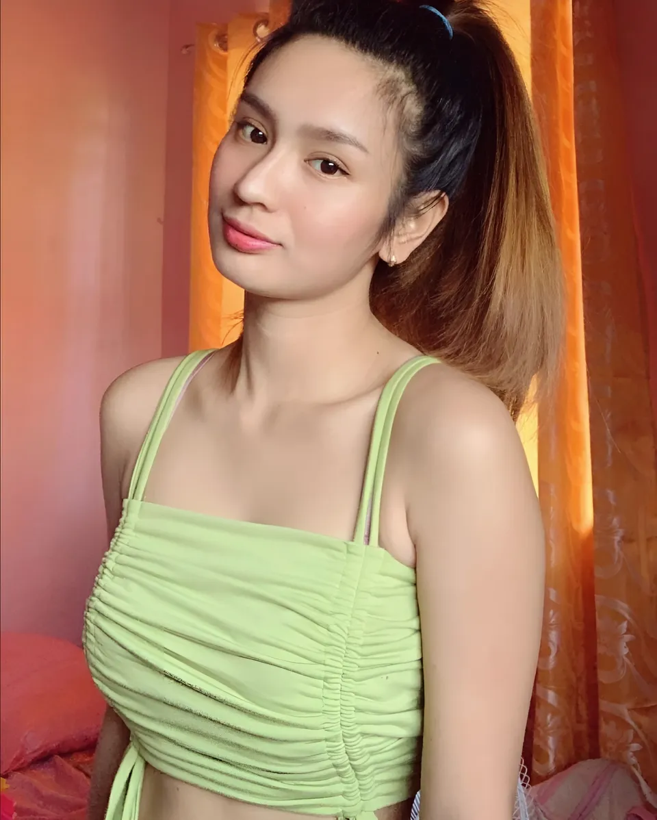 Eunice Lois in green ruched top posing indoors near orange curtain