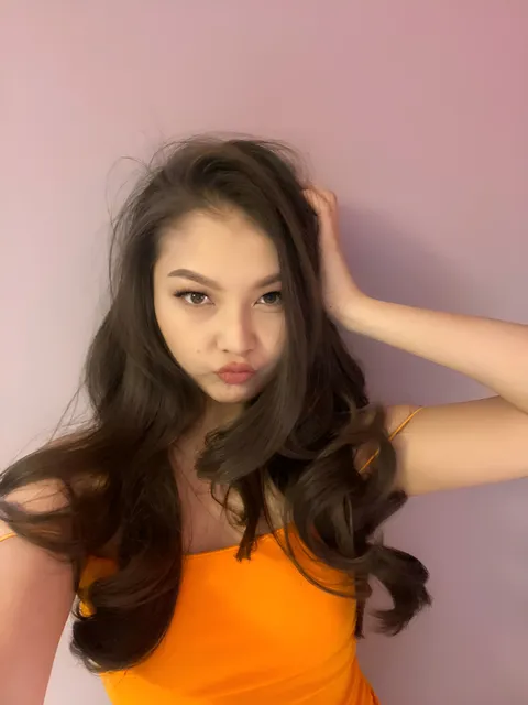 Faith Hartwell in orange top posing with hand in hair against pink wall