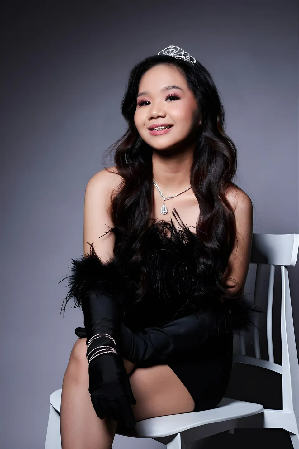 Faye Ramos in black feathered top and gloves seated on white chair
