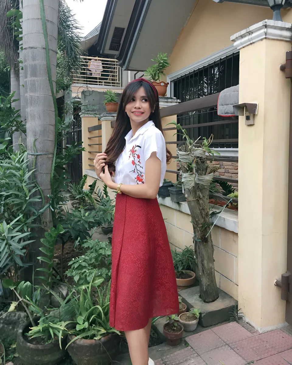 Ica Caparas in red skirt and white top standing outdoors near plants