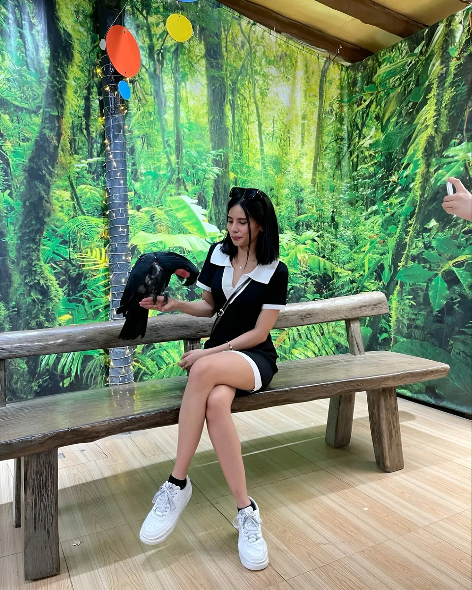Ica Caparas sits on bench with parrot on her hand against jungle mural