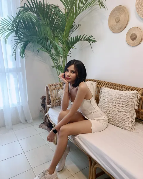 Ica Caparas in white dress seated on wicker bench with plant and pillows