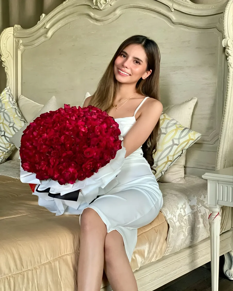 Ica Caparas in white dress holding large red rose bouquet on bed
