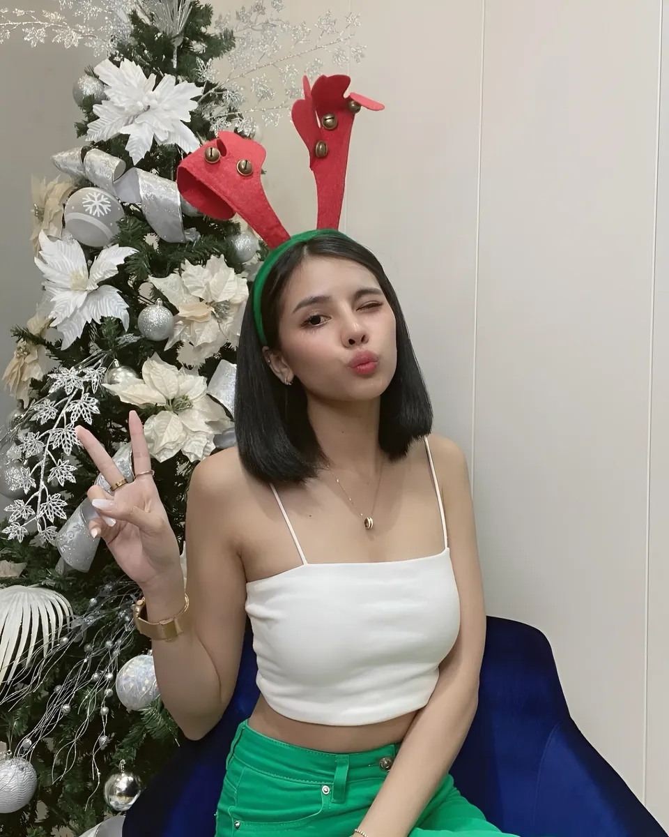 Ica Caparas in white top and green pants seated near Christmas tree