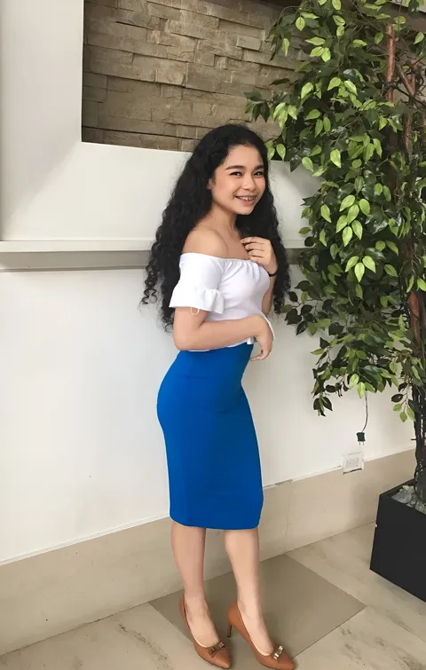 Isla Chelsea wearing a white top and blue skirt poses beside a leafy plant