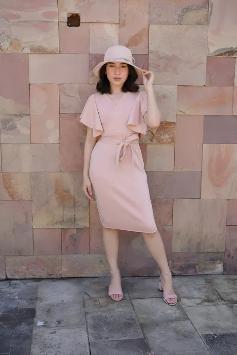 Jewel Hartley in pink dress and hat standing against stone wall