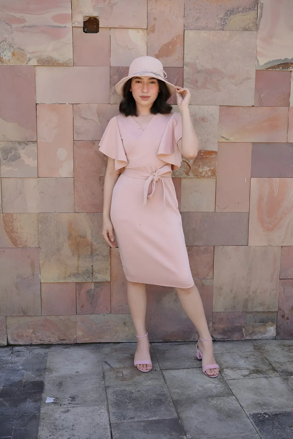 Jewel Hartley in pink dress and hat standing against stone wall