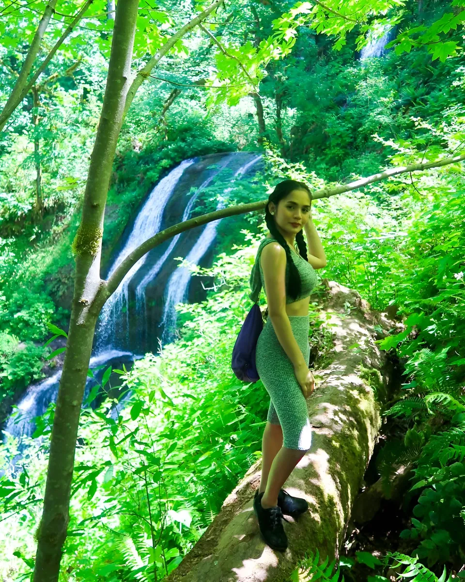 Joey Nerosa in green outfit stands on fallen log near waterfall