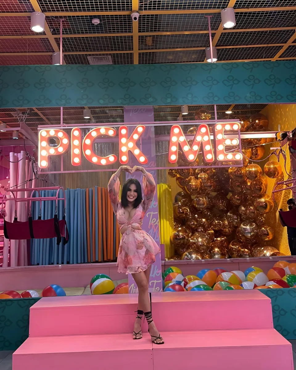 Joey Nerosa in pink dress stands on pink steps under neon sign