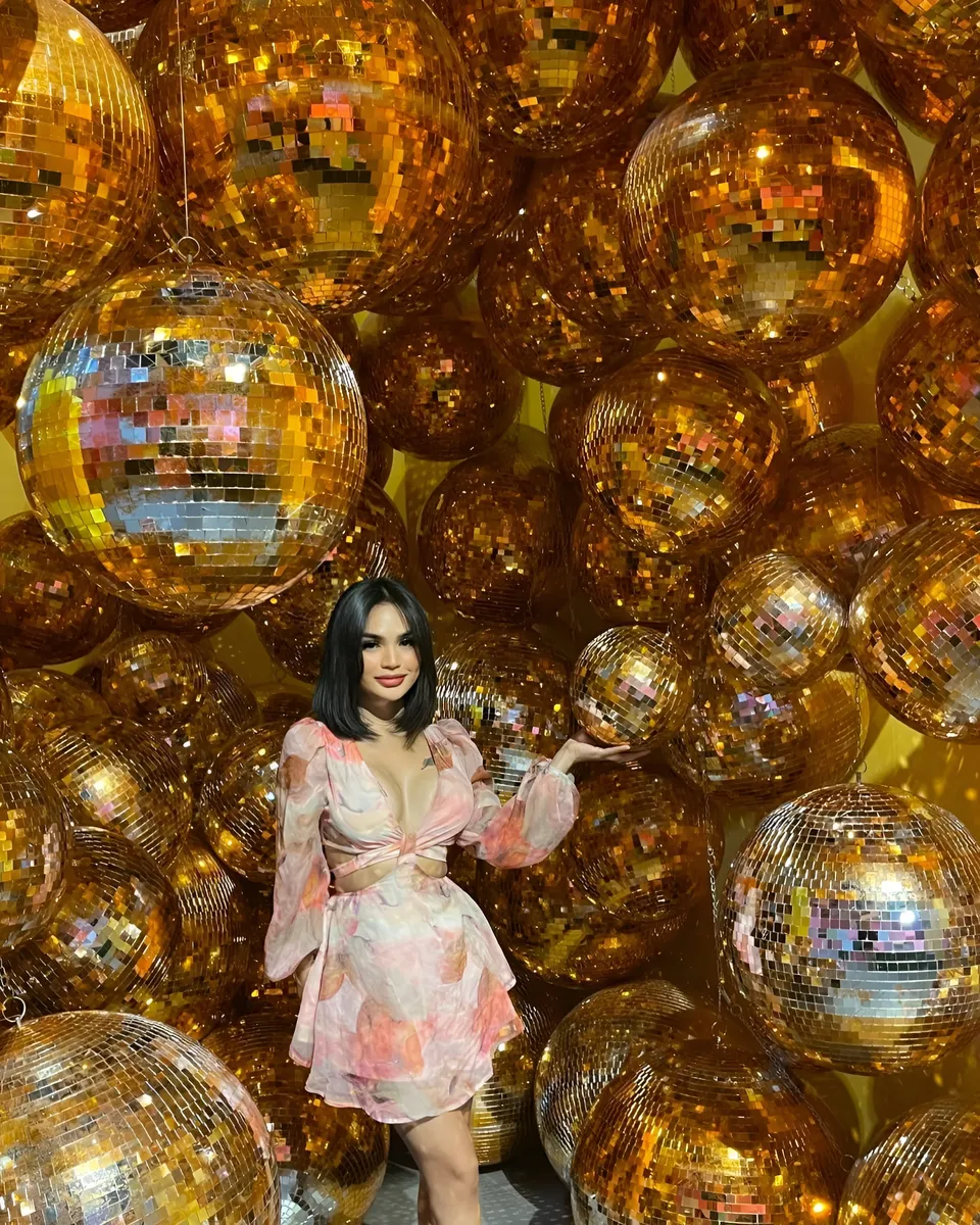 Joey Nerosa in pink floral dress stands among hanging golden disco balls