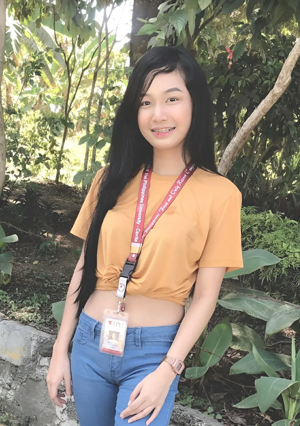 Kara Celeste Pagmel in orange top and blue jeans stands outdoors near foliage