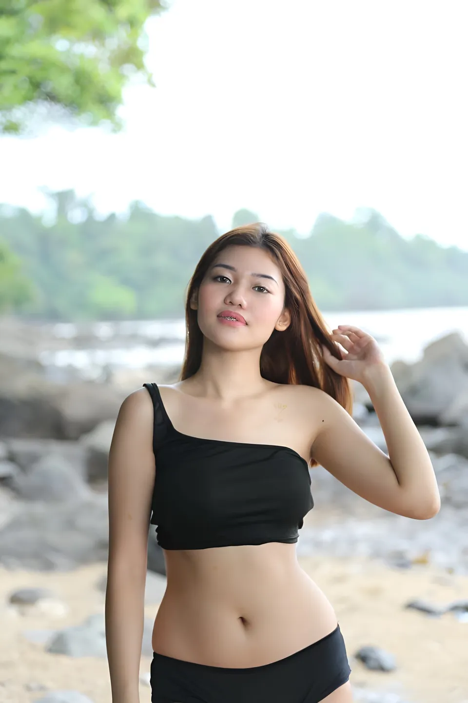 Katrina Alexis in black top standing on beach with rocks and trees