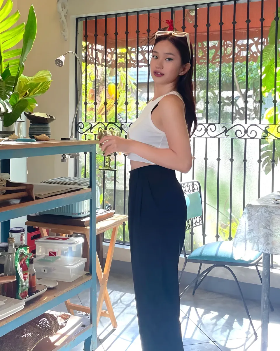 Maeve Linden in white top and dark pants stands near window with plants