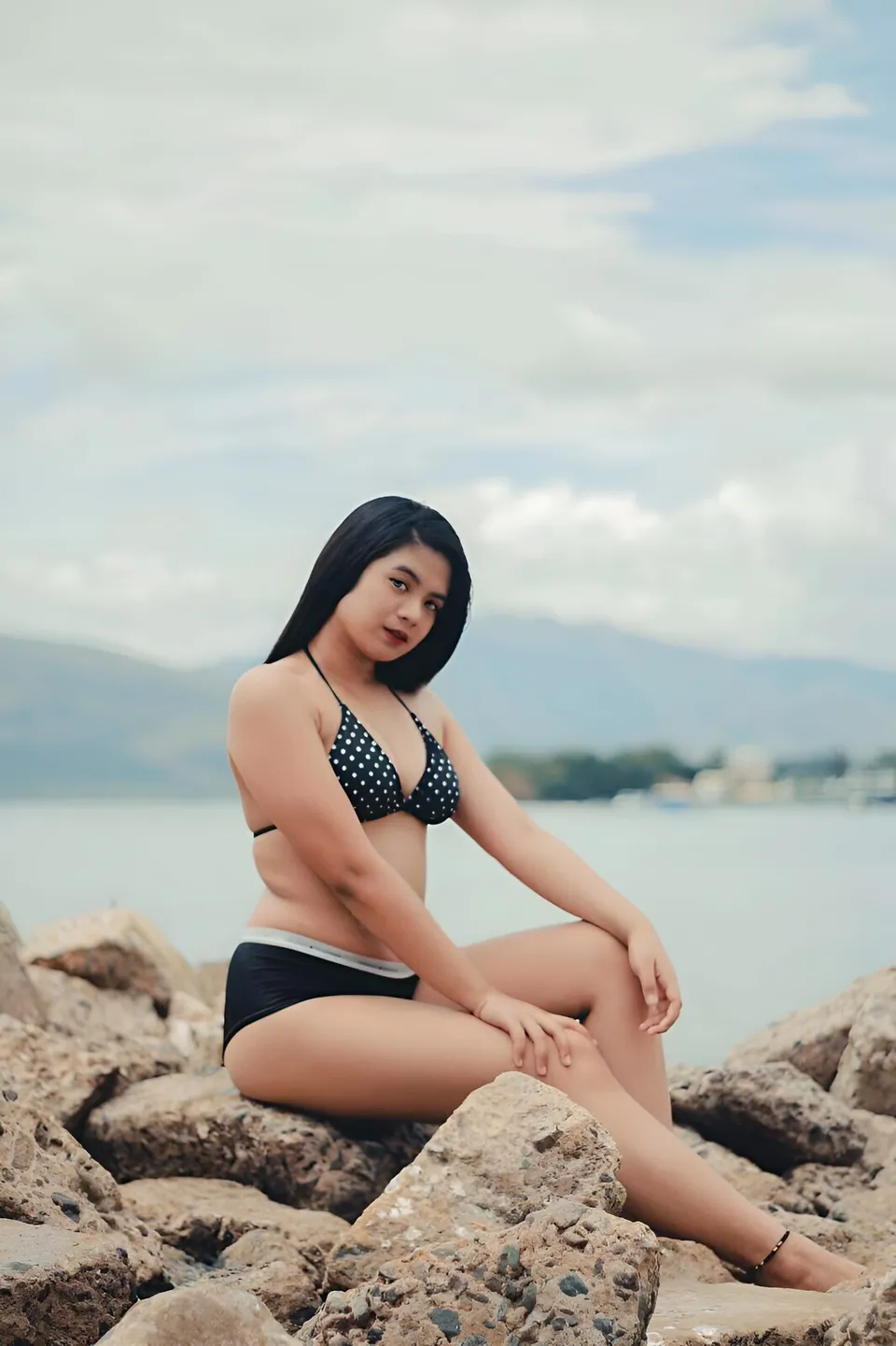 Maria Dacayo in black polka dot bikini sits on rocks by water